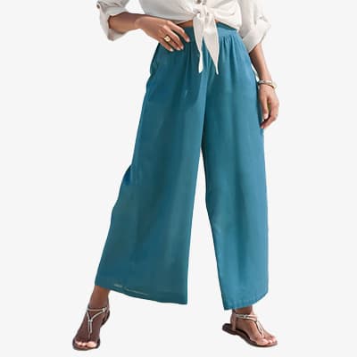 Frau in weiter türkisfarbener Hose, geknoteter Bluse und Sandalen, steht locker mit einer Hand in der Hosentasche