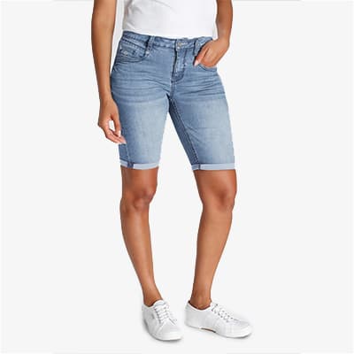 Frau in knielanger, hellblauer Jeansshorts und weißem T-Shirt, steht in weißen Sneakers vor hellem Hintergrund