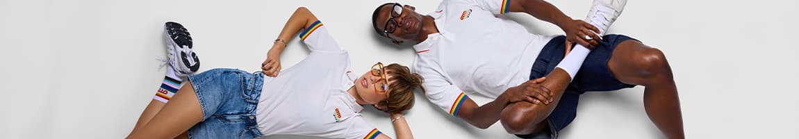 Zwei Personen liegen entspannt auf dem Boden und tragen weiße Poloshirts mit Regenbogen-Details und Shorts