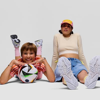 Zwei Personen in Freizeitkleidung posieren entspannt am Boden, eine hält einen Fußball, beide tragen Sportschuhe