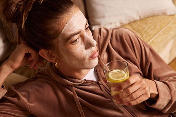 Person im bequemen Hausanzug liegt auf dem Sofa und hält ein Glas Wasser mit Zitronenscheiben in der Hand