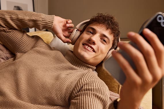 Person in beigem Pullover liegt entspannt auf dem Sofa und schaut auf ein Smartphone