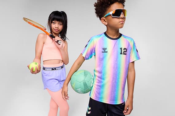 Zwei Kinder in Sportoutfits, Mädchen mit Tennisschläger und Ball, Junge mit Fußball im Regenbogenshirt mit Nummer 12