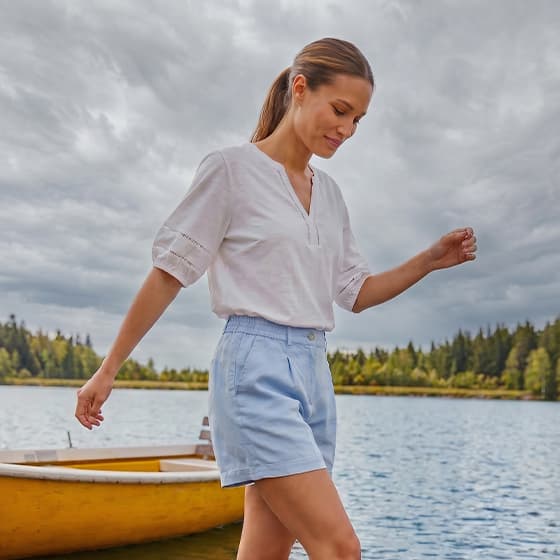 Frau in weißer Bluse und hellblauer Shorts geht am Seeufer neben einem gelben Ruderboot entlang