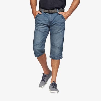 Mann steht mit Händen in den Taschen, trägt knielange Jeansshorts, dunkles Hemd, Gürtel und graue Schnürschuhe
