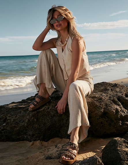 Frau in hellem Sommeroutfit sitzt auf Felsen am Strand und trägt flache Sandalen mit Leo-Muster