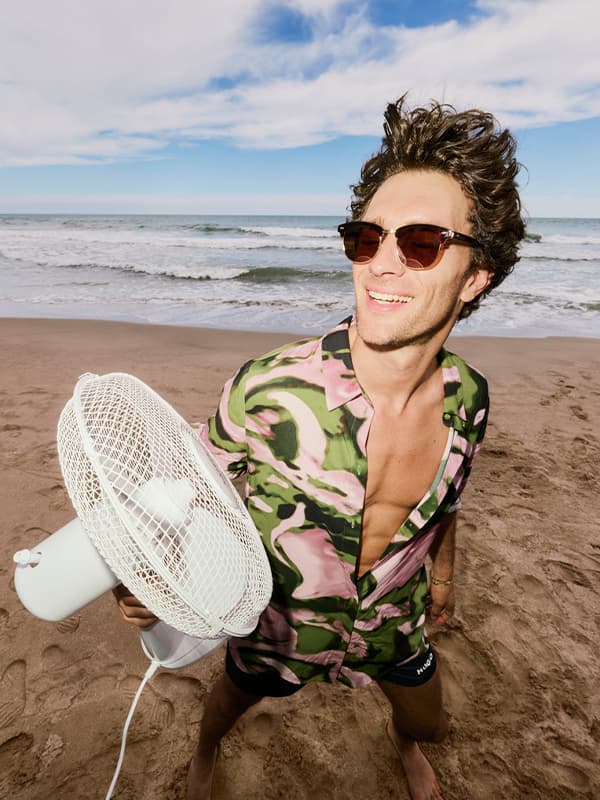 Mann im offenen Camouflagehemd hält einen Standventilator am Strand mit Meer und Wellen im Hintergrund