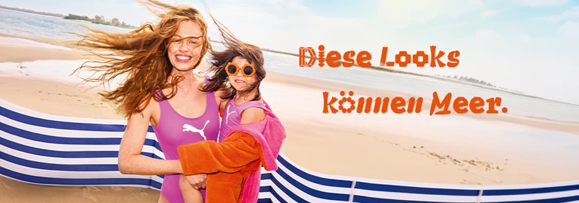 Diese Looks können Meer – Frau im Badeanzug hält Kind im Strandoutfit vor dem Meer