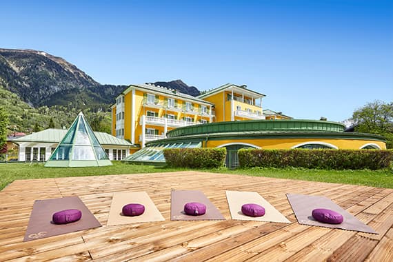 Vier Yogamatten mit lila Kissen auf Holzplattform vor gelbem Hotelgebäude in grüner Berglandschaft