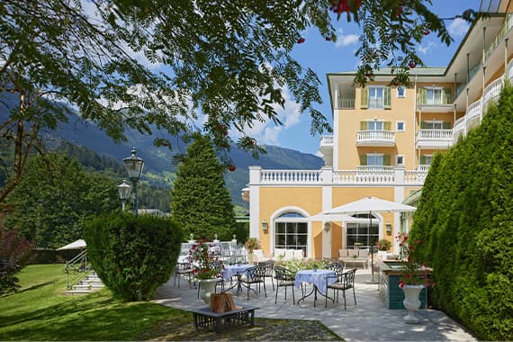 Terrasse eines gelben Hotels mit gedeckten Tischen im Garten, umgeben von Bäumen und Bergen im Hintergrund