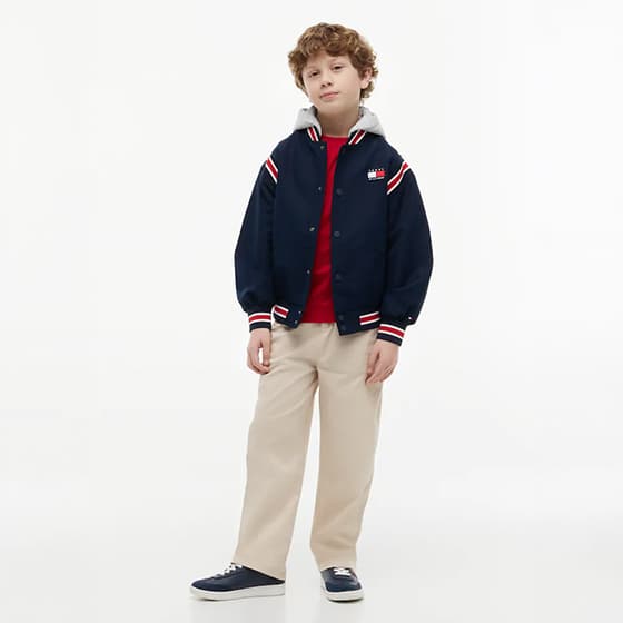 Junge steht locker in navy Collegejacke mit Kapuze, rotem Shirt, beiger Hose und dunklen Sneakern vor weißem Hintergrund