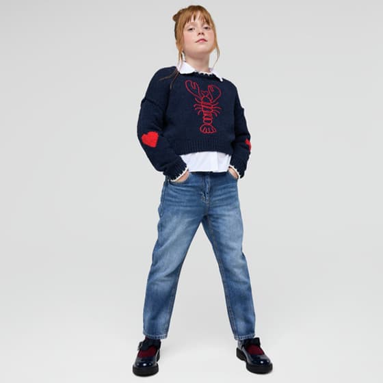 Mädchen steht mit Händen in den Taschen, trägt Jeans und dunklen Pulli mit rotem Hummermotiv und Herz auf den Ärmeln