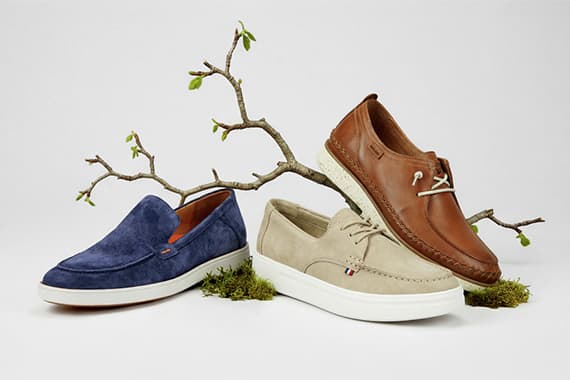 Drei Herren-Slipper in Blau, Beige und Braun stehen dekorativ vor einem Ast mit frischen Knospen