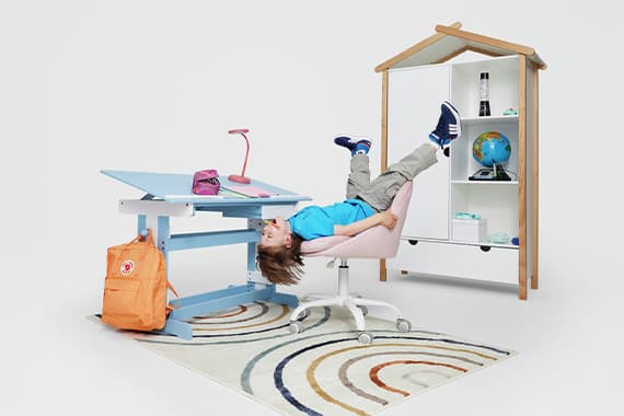 Junge hängt kopfüber auf Schreibtischstuhl zwischen höhenverstellbarem Kinderschreibtisch und Regal in Hausform