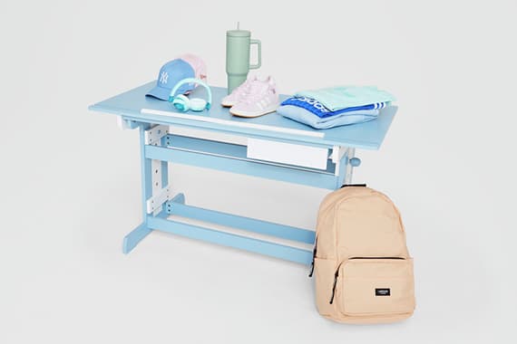 Hellblauer Kinderschreibtisch mit Kappe, Kopfhörern, Trinkbecher, Turnschuhen, Kleidung und beigem Rucksack daneben