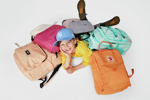 Kind mit blauer Kappe liegt lachend zwischen bunten Rucksäcken auf dem Boden