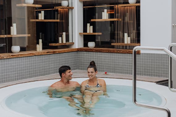 Paar sitzt entspannt im runden Whirlpool und schaut sich an, im Hintergrund Regale mit Kerzen