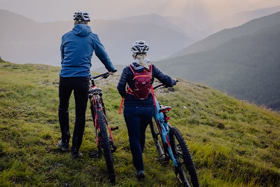 Zwei Personen mit Mountainbikes stehen auf einer Wiese und blicken mit Helmen in ein weites Tal mit Hügeln
