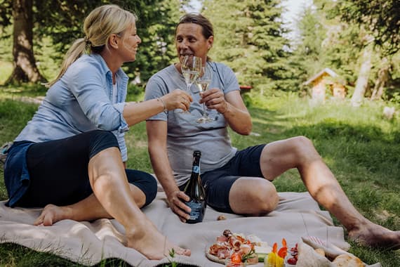 Paar sitzt auf Decke im Grünen, stößt mit Weißwein an, vor ihnen Picknick mit Brot, Käse und Aufschnitt