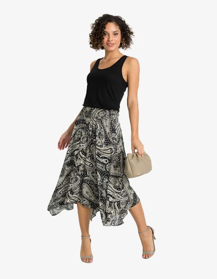 Frau im schwarzen Top und schwingendem Paisley-Midirock, hält beigefarbene Clutch und trägt hohe Sandalen