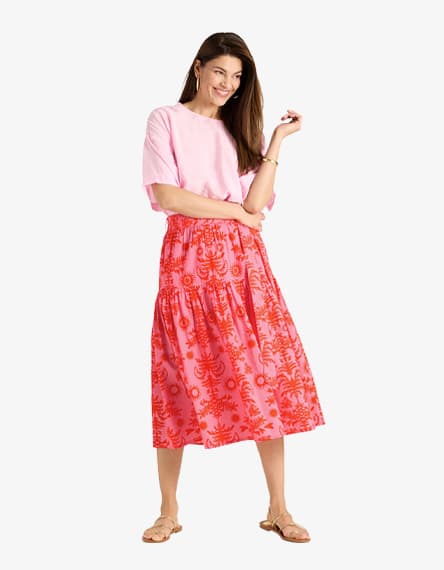 Frau in rosa T-Shirt und pink gemustertem Midirock steht locker und lächelt in die Kamera