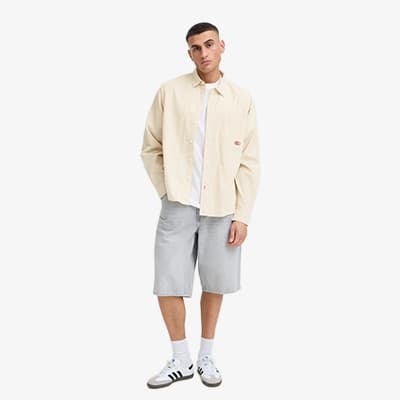 Mann steht leicht schräg in weiter Hemdjacke, T-Shirt, knielangen Shorts, Socken und Sneakers vor weißem Hintergrund