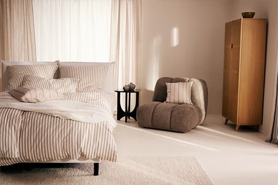 Schlafzimmer mit gestreiftem Bett, beigem Sessel mit Kissen, kleinem runden Beistelltisch und schmalem Holzschrank