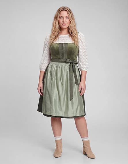 Frau im knieumspielenden grünen Dirndl mit weißer Spitzenbluse und Schürze steht frontal vor grauem Hintergrund