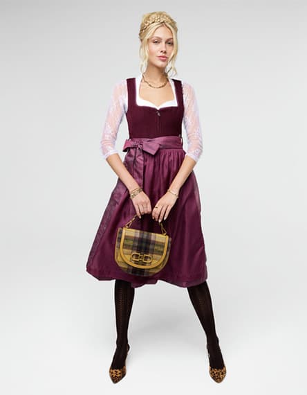 Frau im bordeauxfarbenen Dirndl mit Schleife, hält karierte Handtasche und steht mit leicht gespreizten Beinen vor hellem Hintergrund