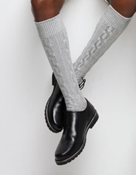 Person mit gekreuzten Beinen trägt kniehohe graue Socken mit Muster und schwarze knöchelhohe Schuhe