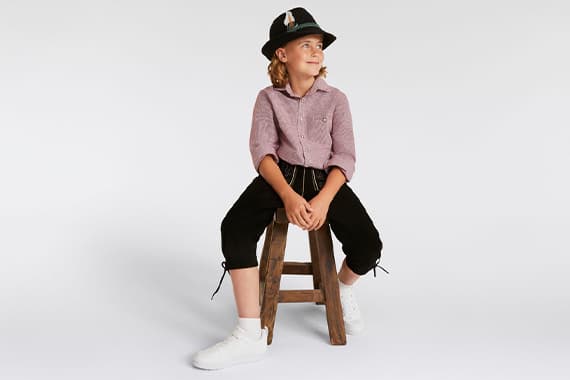 Kind in Trachtenhut sitzt lässig auf Hocker, trägt gemusterte Bluse, knöchellange Hose und weiße Sneaker