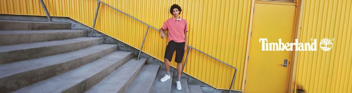 Ein junger Mann steht auf einer Stiege vor einer gelben Wellblechwand und sieht direkt in die Kamera