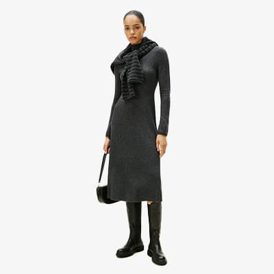 Frau in langem, grauem Kleid mit Schal und schwarzen Stiefeln hält eine schwarze Handtasche