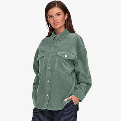 Frau trägt grünes Oversize-Hemd mit Brusttaschen und schwarze, gestreifte Hose, Hände in den Hosentaschen.