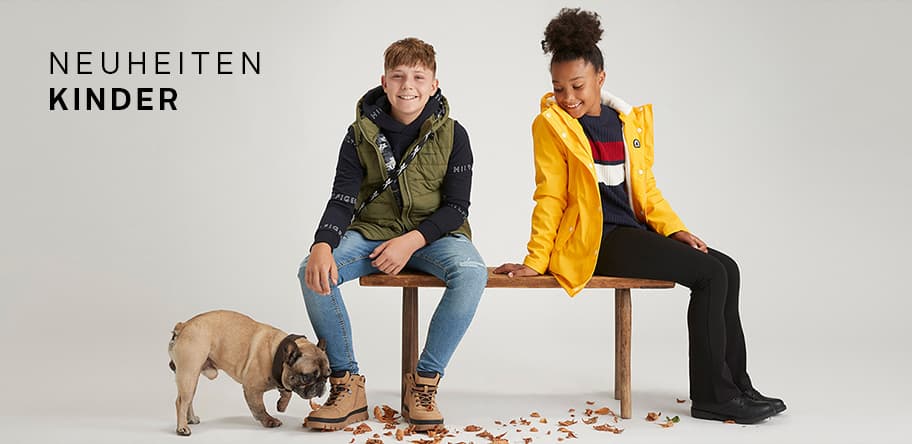 NEUHEITEN KINDER – Junge in grüner Weste und Mädchen in gelber Jacke sitzen auf Bank, Hund schnüffelt am Boden