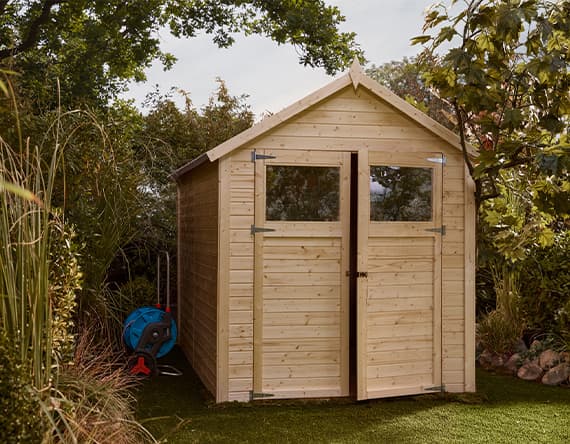 Holzgartenhaus mit zwei Fenstern und Doppeltür, umgeben von Bäumen und Pflanzen, Gartenschlauch auf Spule daneben