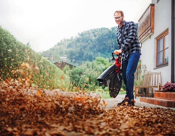 Mann in kariertem Hemd und Jeans bläst Laub mit einem Laubbläser auf einem Gehweg neben einem Haus mit Garten