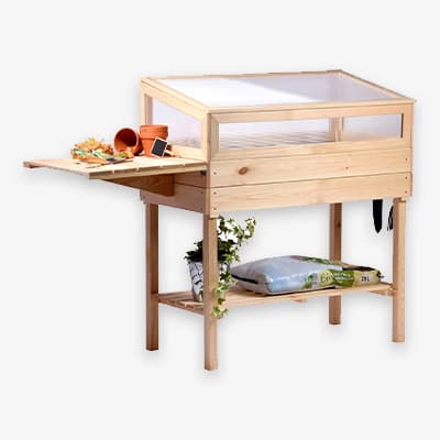 Holz-Gewächshaus mit transparenten Wänden, ausklappbarer Arbeitsfläche und Ablage. Gartenzubehör auf Arbeitsfläche und unterer Ablage.