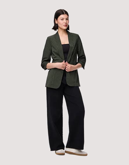 Frau trägt karierten Blazer, schwarzes Oberteil und weite schwarze Hosen. Hände halten Blazer vorne zusammen.