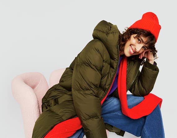 Frau in langer, olivgrüner Daunenjacke und roter Mütze sitzt auf rosa Sessel, trägt rote Schal und blaue Jeans