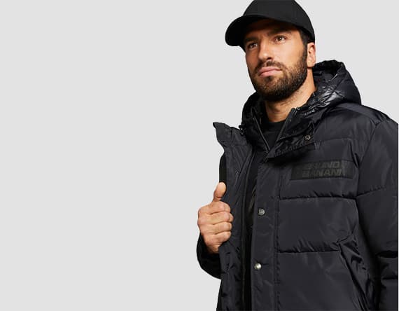 Mann trägt schwarze Winterjacke mit Kapuze und Stehkragen. Er hält die Jacke am Kragen fest und trägt eine schwarze Baseballkappe.