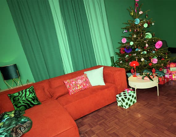 Wohnzimmer mit rotem Sofa, grünen Kissen, Weihnachtsbaum mit buntem Schmuck, Geschenke auf dem Boden, grüne Vorhänge im Hintergrund
