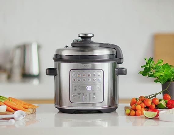 Elektrischer Multikocher auf Küchenarbeitsplatte, umgeben von frischen Karotten, Tomaten und Kräutern.