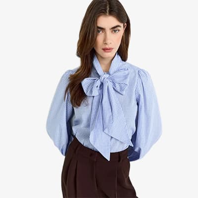 Frau trägt eine blau-weiß karierte Bluse mit großer Schleife und brauner Hose.