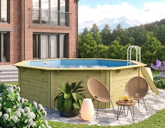 Runder Holzpool im Garten vor Backsteinhaus, umgeben von Pflanzen und Gartenmöbeln mit Bergblick im Hintergrund