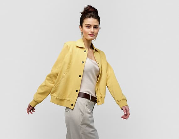 Frau trägt gelbe Jacke, weißes Top und beige Hose, posiert mit ausgestreckten Armen.