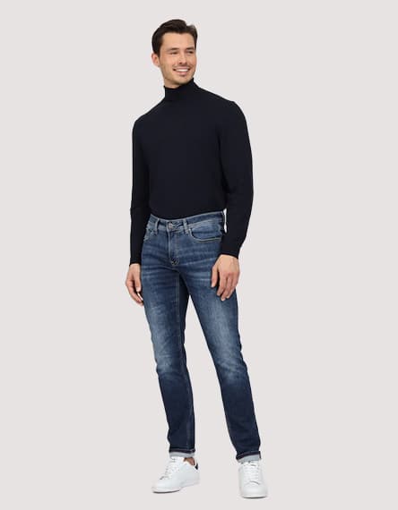 Mann in dunkelblauem Pullover und blauen Jeans, trägt weiße Sneakers.