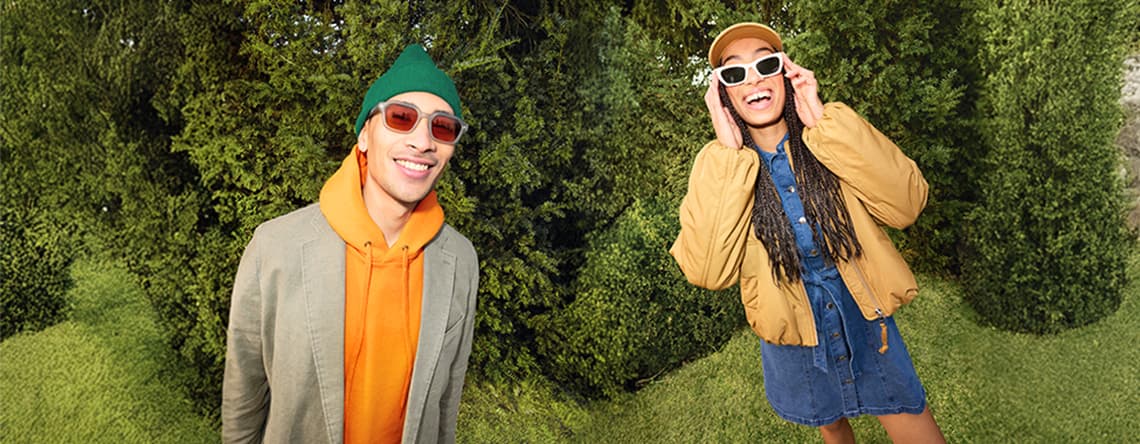 Zwei Personen in lässiger Outdoor-Mode stehen vor dichtem Grün, links Hoodie und Blazer, rechts Jeanskleid mit Jacke