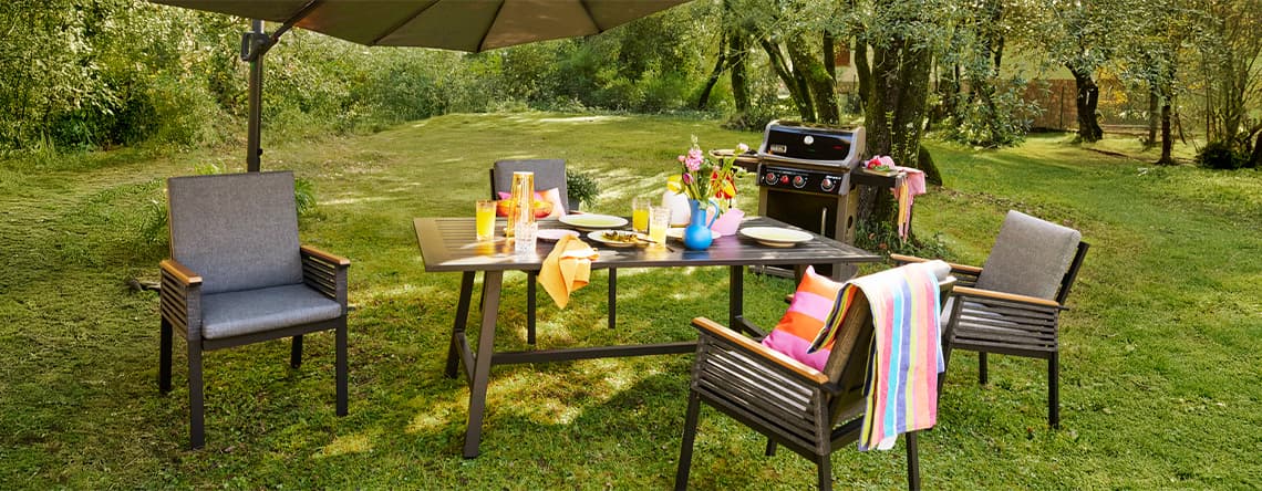 Gartentisch mit vier Stühlen, bunten Gläsern und Handtüchern, daneben Grill unter großem Sonnenschirm auf Rasenfläche