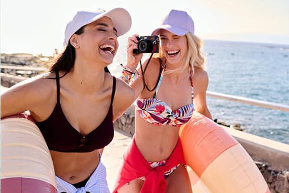 Zwei Frauen in Bikinis mit Schwimmringen am Meer, eine hält eine Kamera und fotografiert Richtung Betrachter
